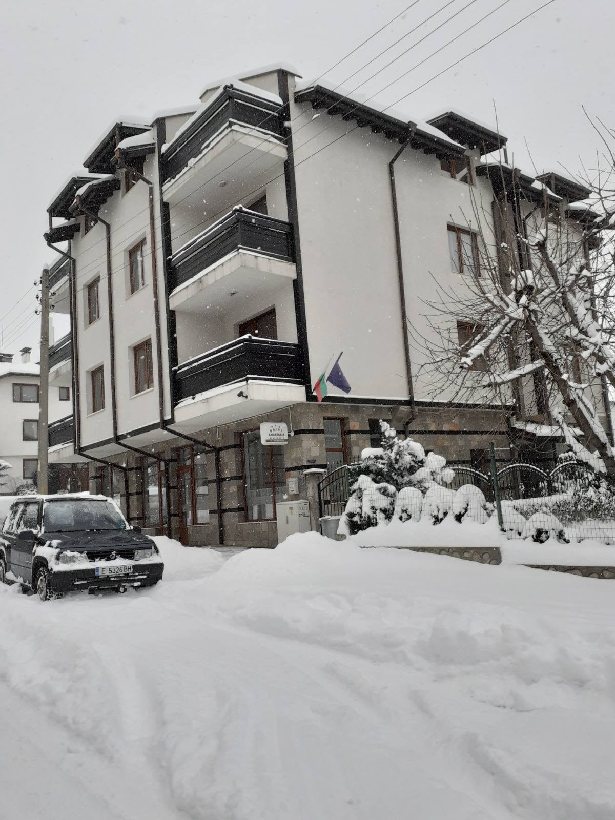 A modern three-story building is shown, partially covered in snow. Black balconies adorn the facade, and a vehicle is parked in front. Snow blankets the ground and trees, creating a serene winter scene. Flags are visible near the entrance.
