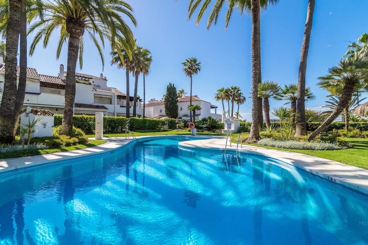 A tranquil swimming pool is surrounded by palm trees and well-kept gardens. Bright blue water reflects the clear sky, while lounge chairs are positioned along the pool's edge. Surrounding greenery adds a lush feel to the outdoor space.