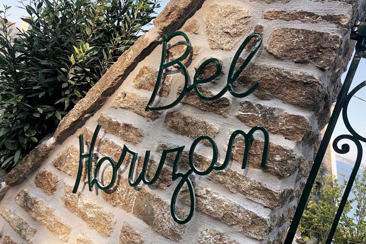 A stone wall displays the name 'Bel Horizon' in green cursive lettering, surrounded by lush greenery. The natural textures of the stones contrast with the smooth metal of the sign, creating an inviting entrance to the villa.