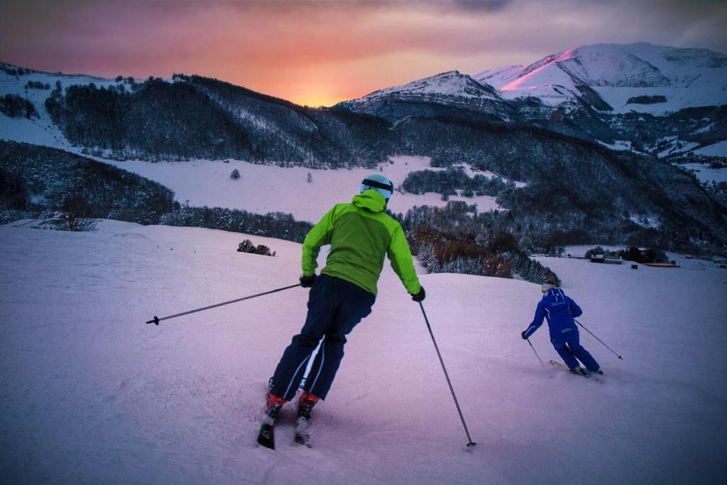 Two skiers are seen descending a snow-covered slope, surrounded by mountainous terrain. The landscape features a gradient sky transitioning into hues of pink and orange, indicating sunrise or sunset. Snow-covered hills create a tranquil backdrop, while the skiers are dressed in colorful winter attire.
