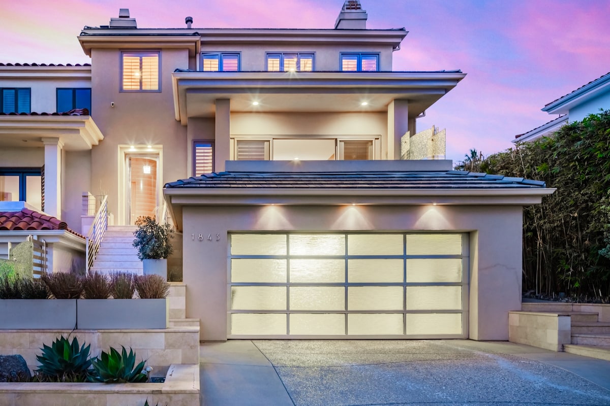 A modern coastal home features a prominent two-car garage with a sleek glass door. The structure displays neutral colors and architectural details under a vibrant sky at dusk. Lush landscaping complements the entrance, leading to a welcoming staircase.