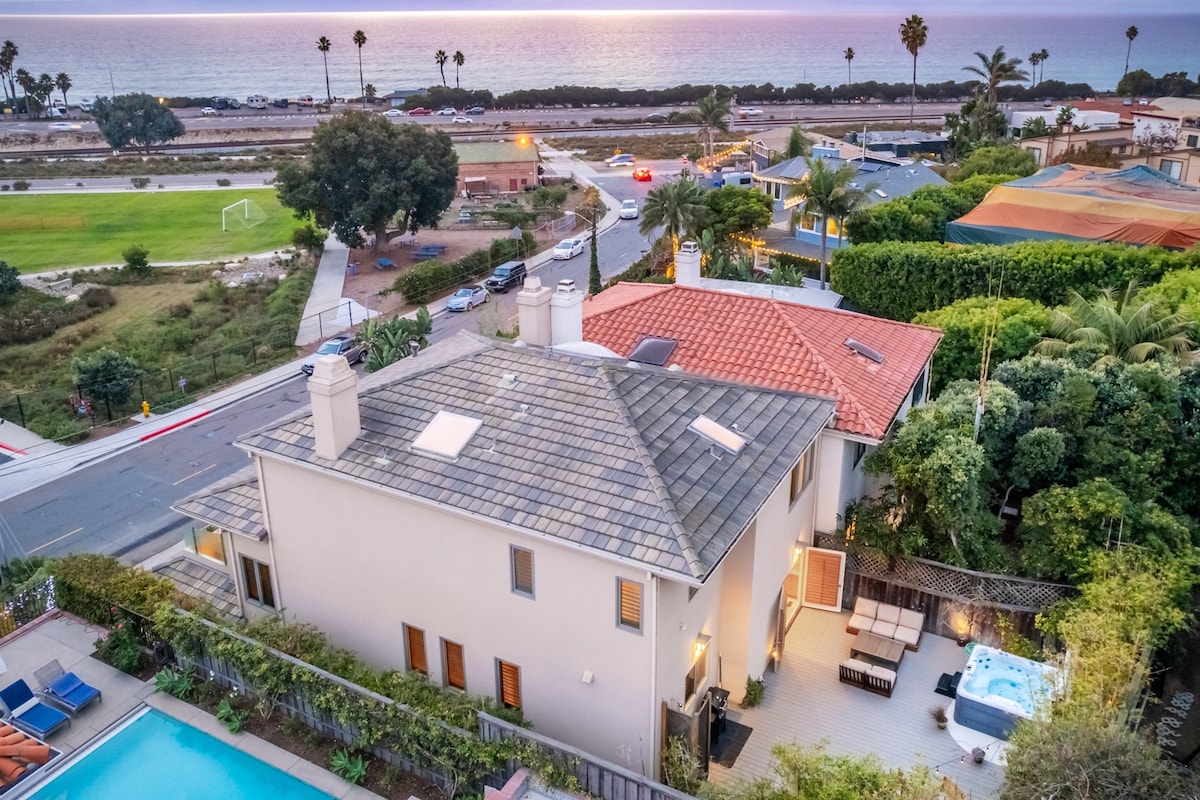 An aerial view captures a modern coastal home positioned near the shoreline, showcasing a beautifully landscaped yard. A private jacuzzi tub sits adjacent to a sitting area, while the ocean reflects the evening light. Palm trees and other greenery enhance the tranquil seaside setting.