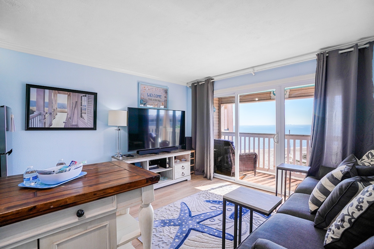 A cozy living room is presented, featuring a dark sofa with decorative pillows. A large television is positioned on a console table. Sunlight illuminates the space, showcasing framed artwork and a basket of snacks. Sliding glass doors lead to a balcony with a beach view.