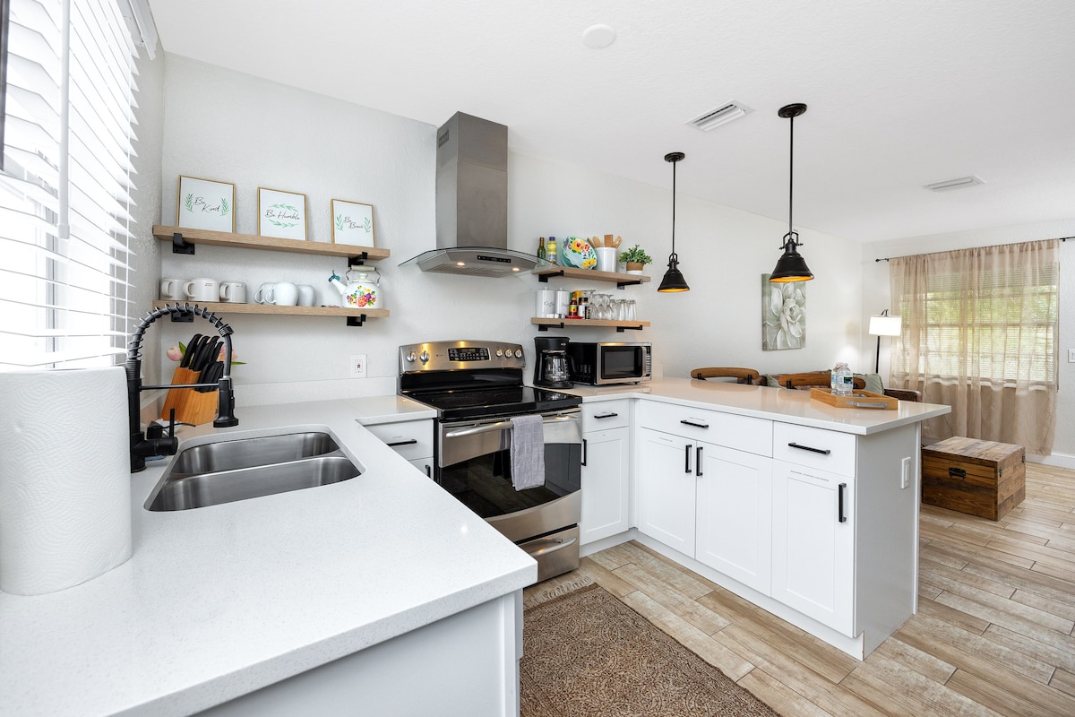 The kitchen features modern appliances, including a stove and microwave, along with ample counter space and open shelves holding dishes and cooking essentials. A sink is positioned beneath a window, while light streams into the room, creating a bright and functional cooking area.