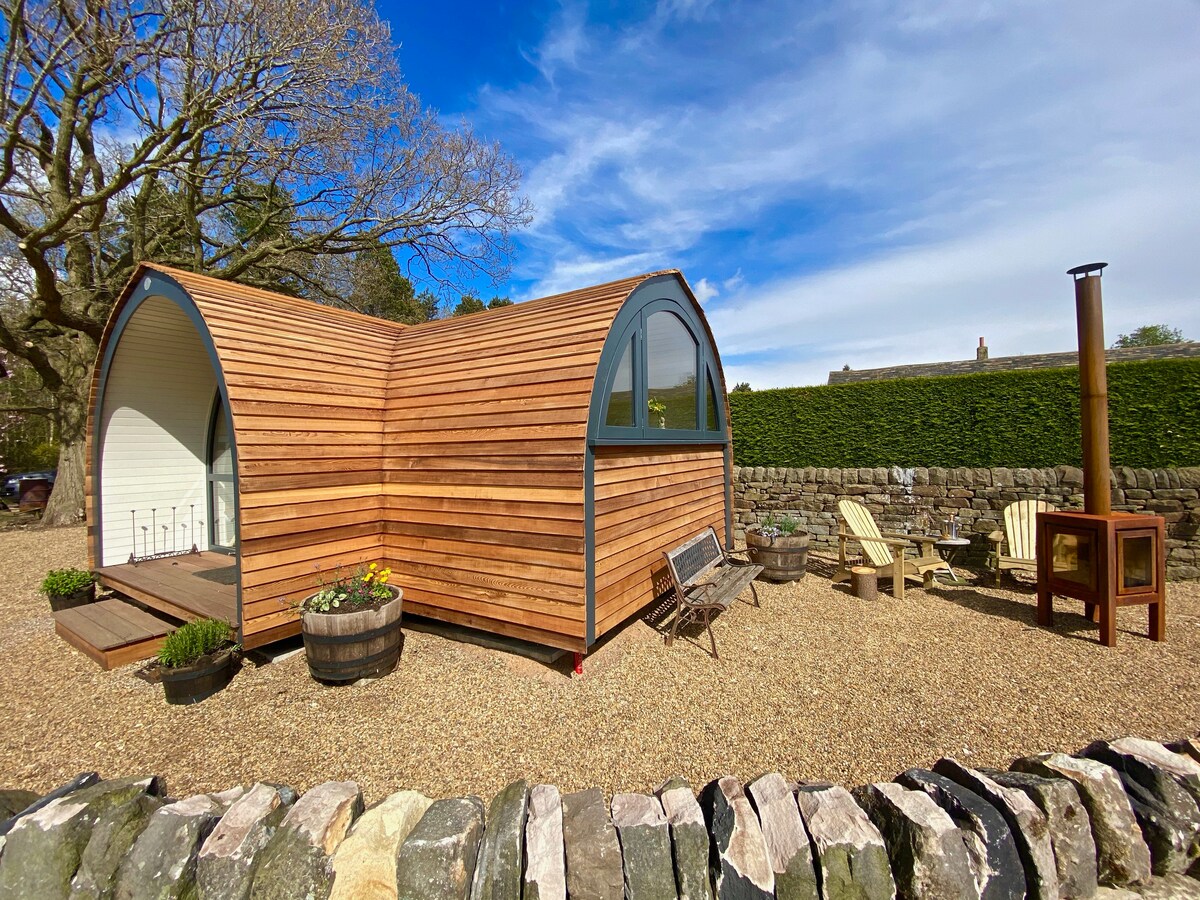 A cabin shaped structure with a wooden exterior is surrounded by gravel and garden. Outdoor seating options include wooden chairs and a table, accompanied by decorative planters. A fireplace is positioned nearby for warmth during cooler evenings.