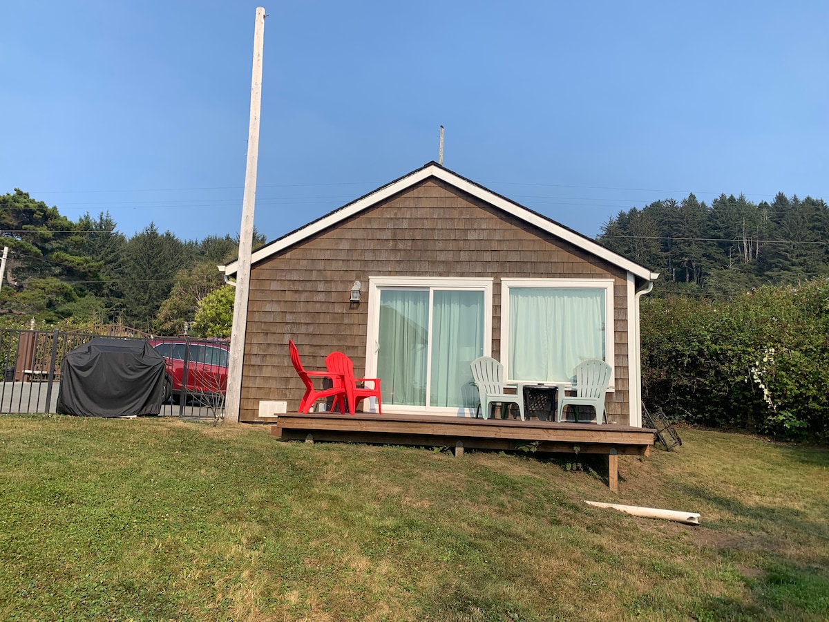 A separate studio guest cottage is depicted, featuring large sliding glass doors that lead to a small deck area. Two lightweight chairs and a small table are situated on the deck, surrounded by a grassy lawn. The property is framed by trees in the background.