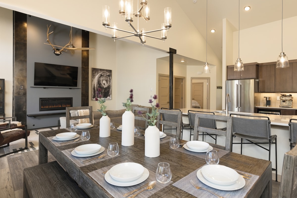 A large dining table is centrally positioned beneath a stylish chandelier, set with plates and glassware. A modern kitchen area is visible in the background, featuring stainless steel appliances. Warm tones and natural textures highlight the inviting atmosphere of the open living space.