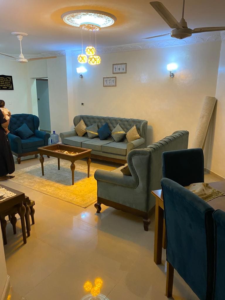 A comfortable living area features a light-colored tiled floor and cream walls, with a soft area rug beneath a wooden coffee table. Two tufted sofas in teal are positioned along with decorative cushions, while ambient lighting is provided by stylish hanging fixtures and wall-mounted lights.