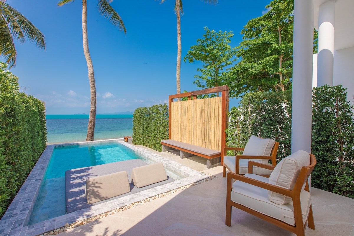 A serene outdoor space features a private pool surrounded by lush greenery and swaying palm trees. Comfortable lounge chairs lie beside the pool, offering a tranquil setting with unobstructed views of the sea and clear blue sky.
