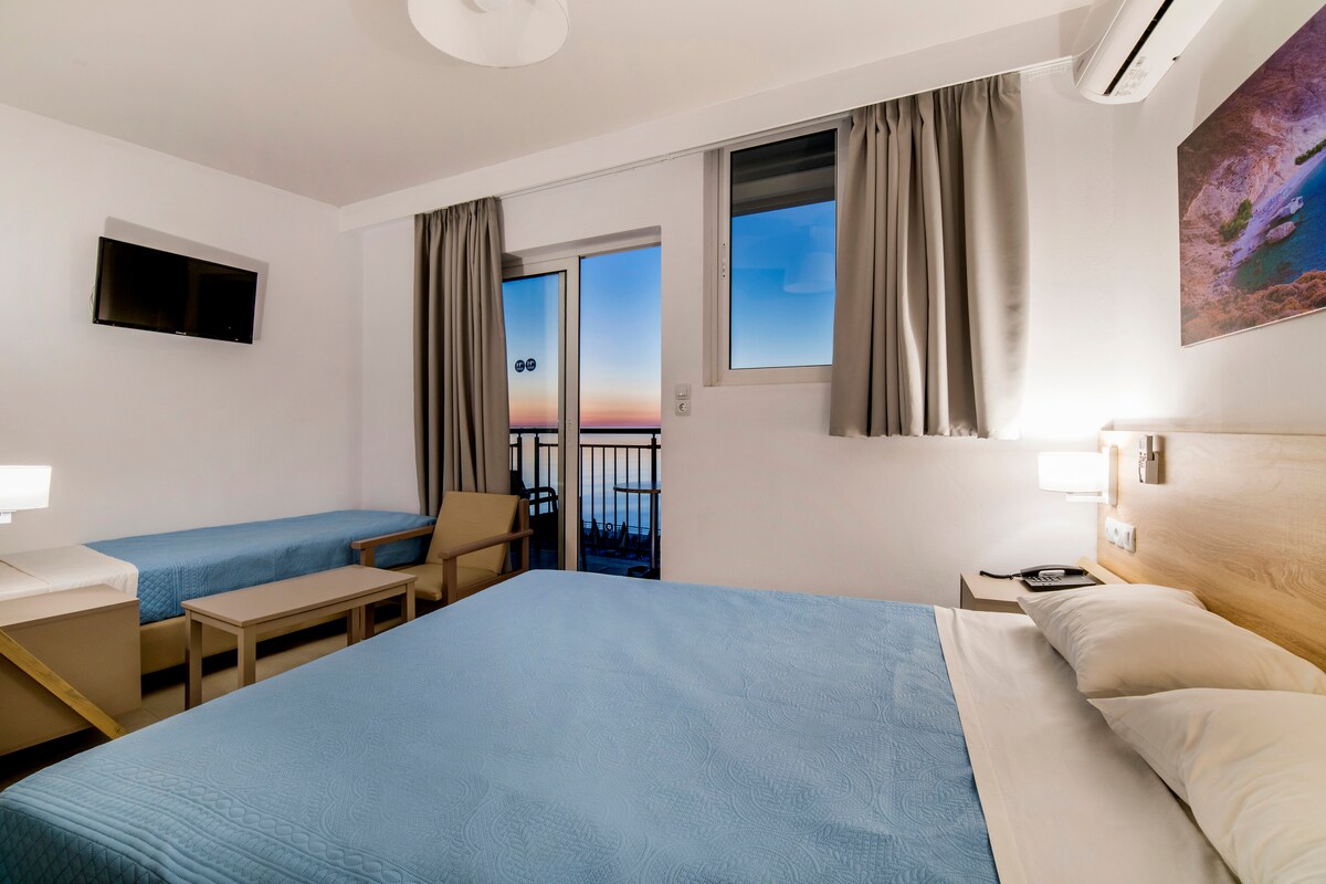 A modern guest room is furnished with a spacious bed and a sofa bed, complemented by a small table and armchair. Large windows offer views of the colorful sky at sunset, while light-colored curtains frame the windows. A flat-screen TV is mounted on the wall.
