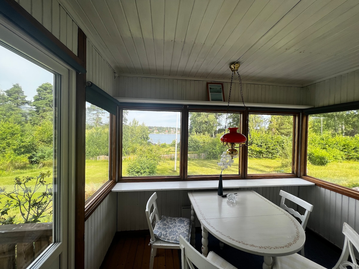 A sunlit sunroom is framed by large windows, offering a view of lush greenery and a serene lake. A white dining table is surrounded by four white chairs, and a red pendant light adds a pop of color to the space.
