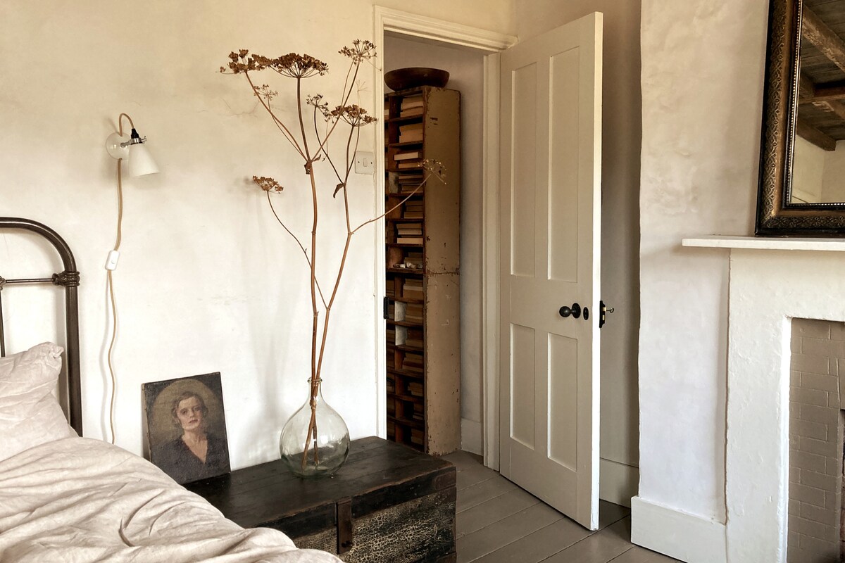 A serene bedroom features a vintage iron bed with soft linen bedding. A tall dried floral arrangement in a glass vase stands beside a dark-stained nightstand. A doorway leads to an adjacent room filled with books, while light illuminates the softly textured wall.