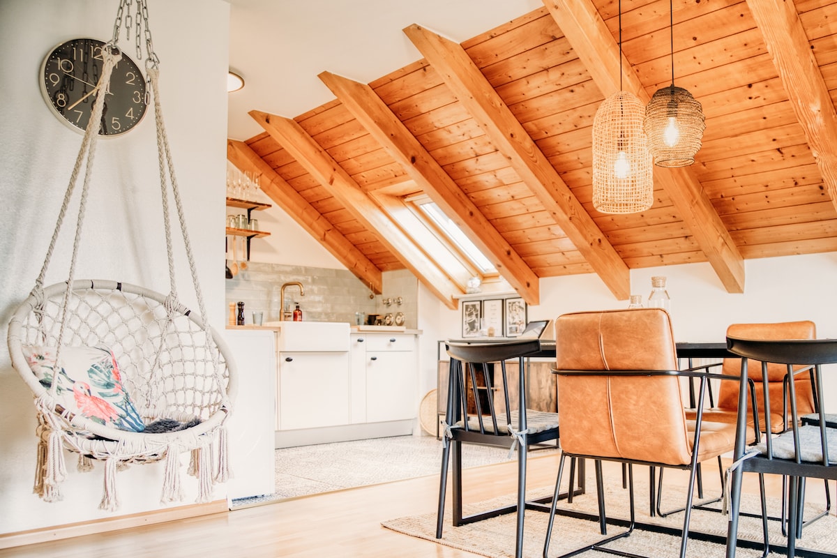 An open living and dining area is distinguished by sloped wooden ceilings and natural light streaming through skylights. A stylish hanging chair adds a cozy touch, while a dark dining table with four chairs invites communal meals. The kitchen area, visible in the background, features modern appliances and a neutral color palette.