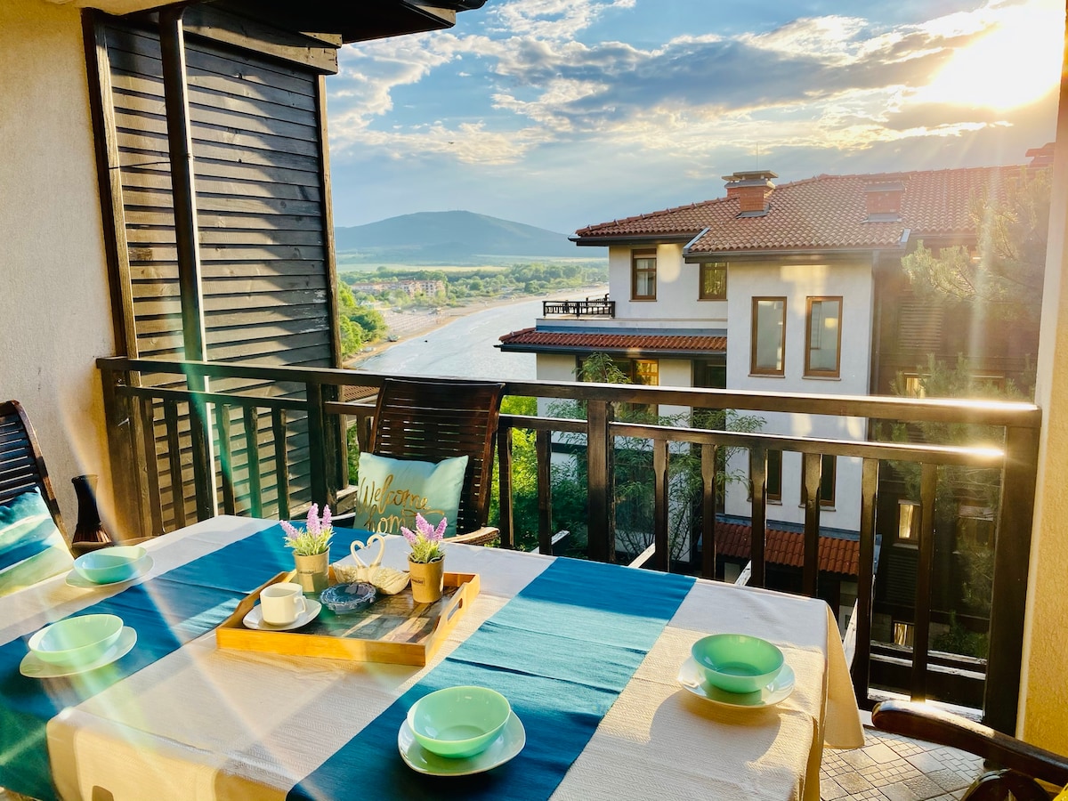 A terrace setting features a dining table adorned with a blue and cream tablecloth, complete with plates and a teacup. Lush greenery and scenic views of the surrounding landscape are visible, while the sun casts a warm glow over the area, highlighting the natural beauty.