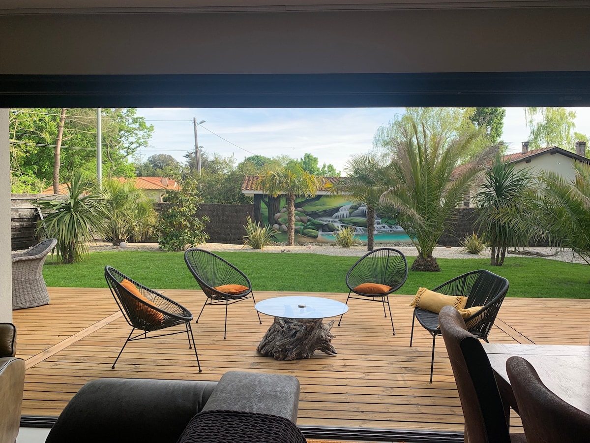 An outdoor terrace showcases a wooden deck with a round table positioned at the center. Surrounding armchairs with cushions invite relaxation. Lush greenery and palm trees provide a serene backdrop, complemented by a decorative mural of a landscape in the distance.