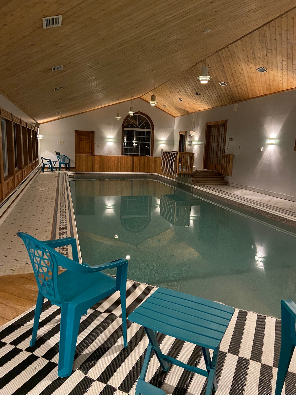 An indoor pool area features calm water under soft lighting. Aqua-colored chairs and a small table sit on a striped rug beside the pool, enhancing the space's functionality. Wooden accents highlight the ceiling and walls, contributing to a warm environment.