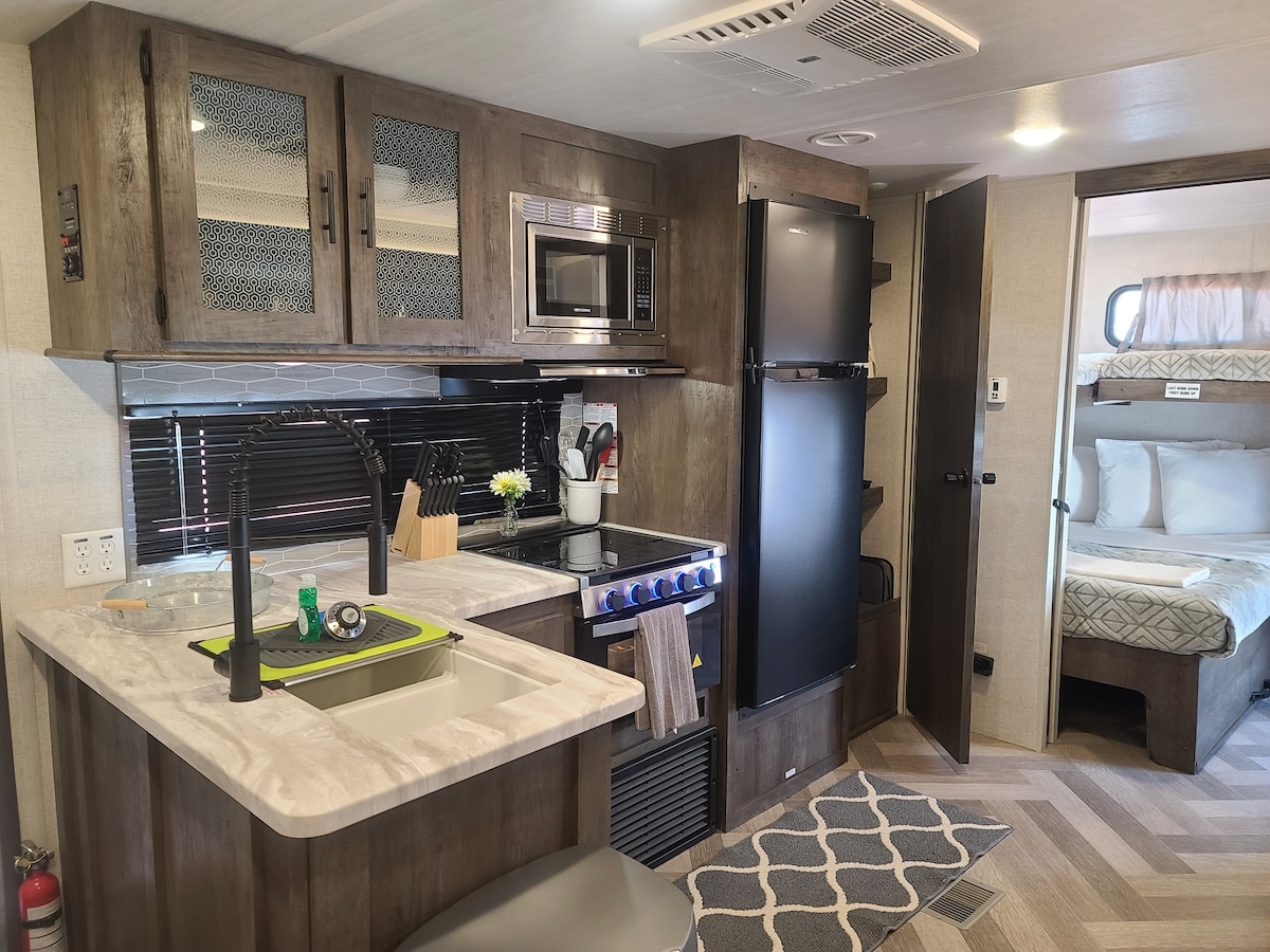 A well-equipped kitchenette features modern appliances, including a microwave and refrigerator. Stylish cabinets provide ample storage, while a sink with a cutting board showcases functionality. A small rug anchors the space, leading into a comfortable sleeping area visible in the background.