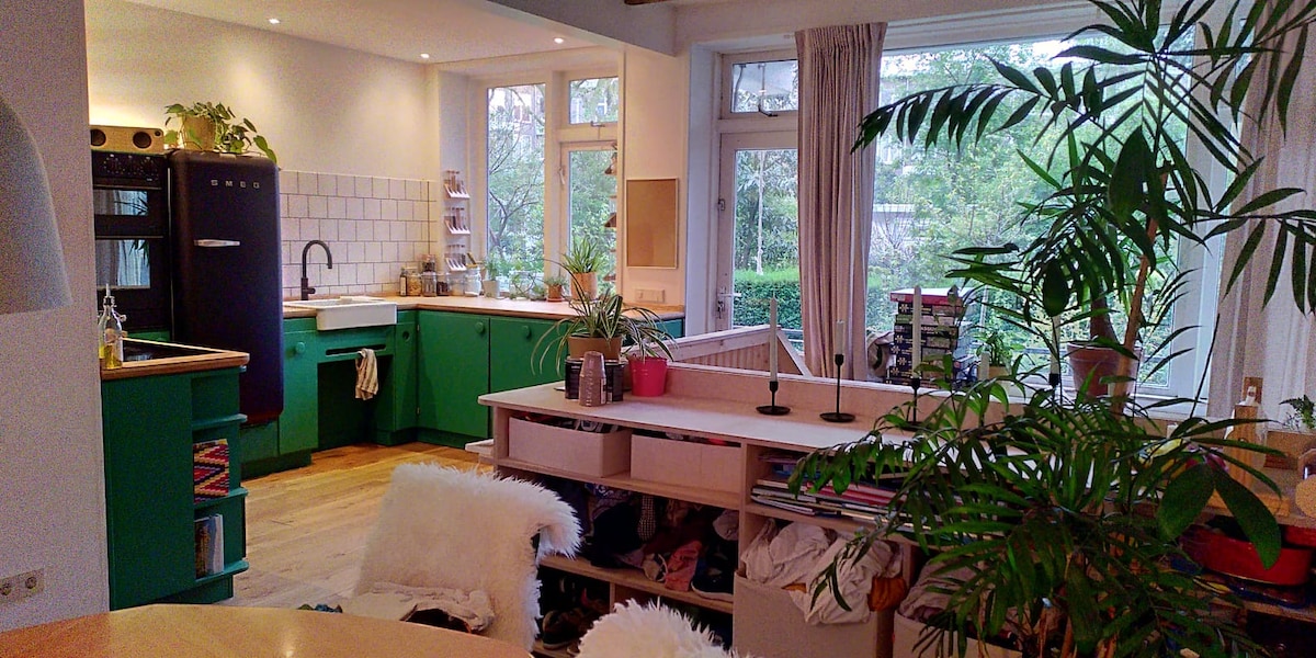 The spacious kitchen features green cabinets and a large sink, complemented by a wooden countertop. Natural light filters through multiple windows, illuminating various indoor plants. An organized shelving unit and a dining table with white fluffy chairs are also visible in the welcoming environment.