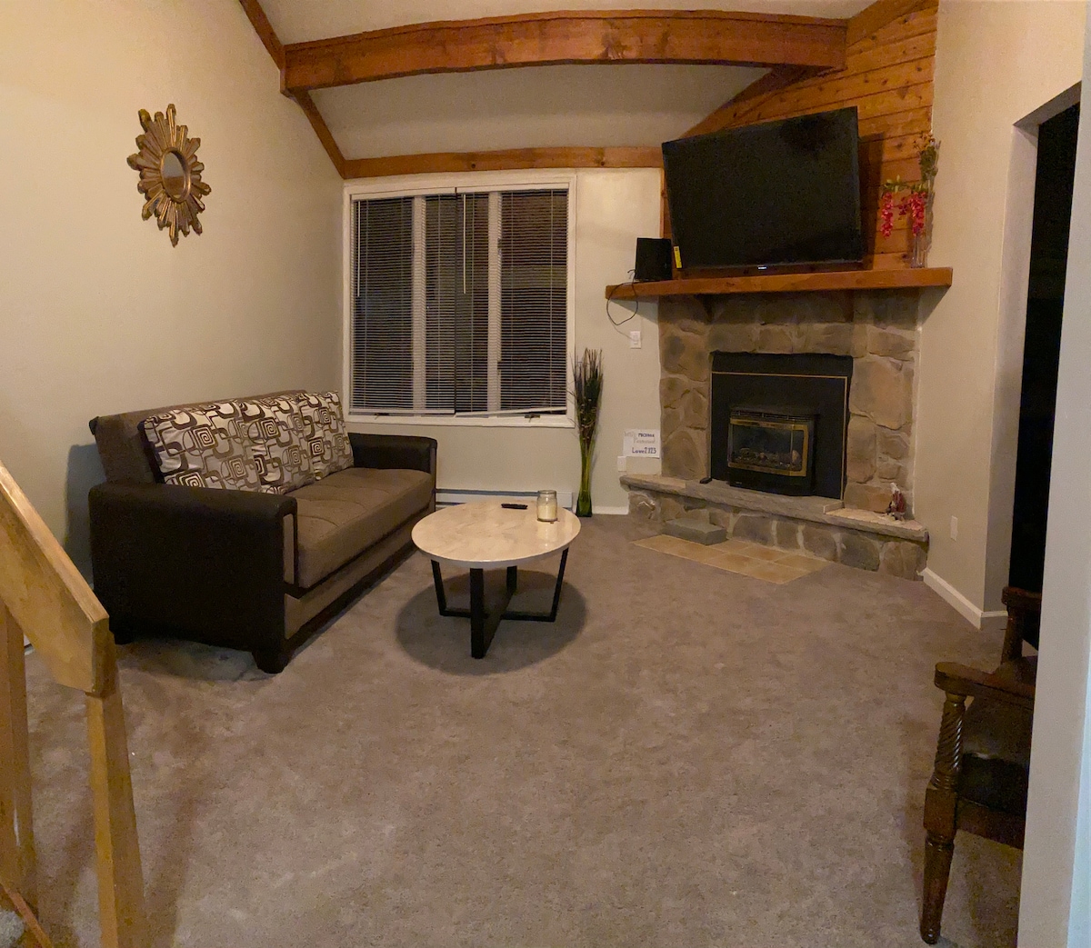 A cozy living area features a comfortable sofa set against a stone fireplace. A round coffee table rests in the center, complemented by a mounted television above the mantel. Light streams through the large window, accentuating the warm wooden beams overhead.