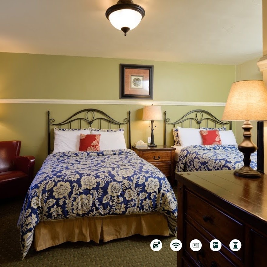 The room features two extra-long Full Size beds with floral-patterned covers, complemented by warm bedside lamps. A dark wooden nightstand is positioned between the beds, and a cozy red armchair is present in the corner, enhancing the room's welcoming atmosphere.