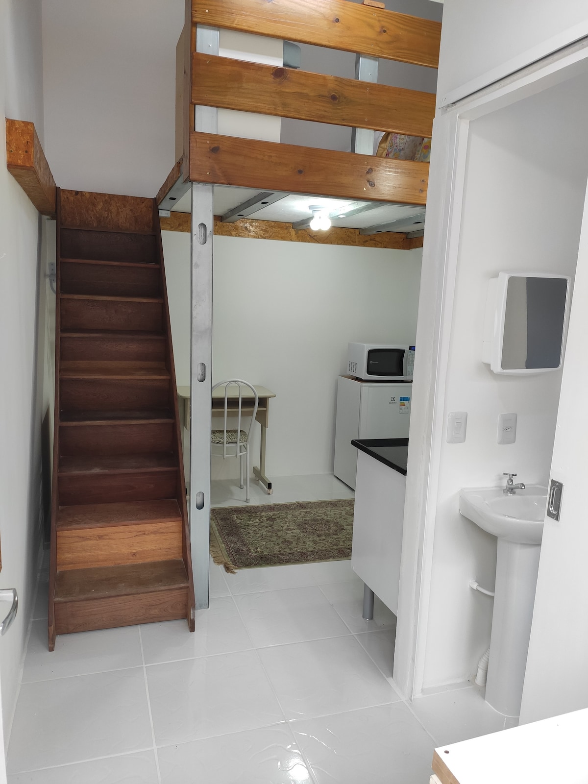 A wooden staircase leads to a small mezanine, with a cozy space for sleeping. The lower area contains a practical kitchenette with a microwave, refrigerator, and sink, alongside a simple table and chair set. A compact bathroom is visible to the right.