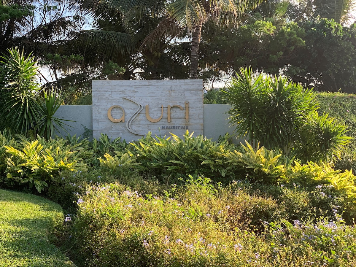 The entrance sign of Azuri is prominently displayed, surrounded by lush tropical vegetation and colorful flowering plants. The sign is designed with a modern font, highlighting the name 'Azuri' against a backdrop of green foliage and palm trees.