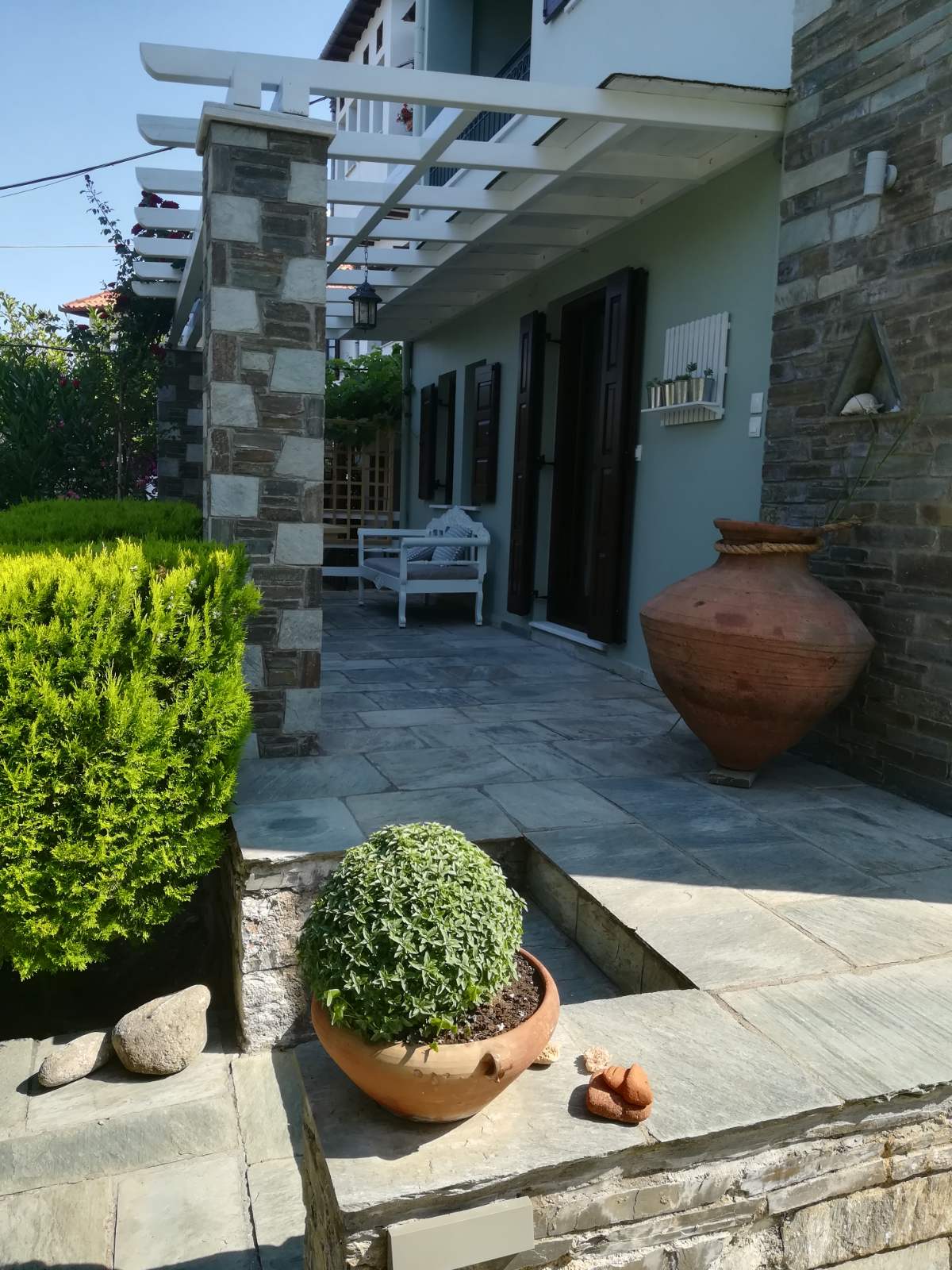 A stone pathway leads to the entrance of a modern building, flanked by manicured greenery and decorative pottery. A terracotta pot holds a small plant, and smooth stones are presented on the pathway, creating an inviting approach to the property.