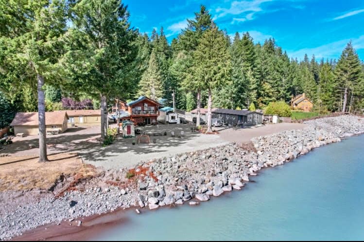 The property showcases a riverside setting surrounded by tall evergreen trees. Several structures, including a cozy main house and additional accommodations, are positioned along the shoreline. The rocky waterfront leads to clear water, with a spacious area for outdoor activities.