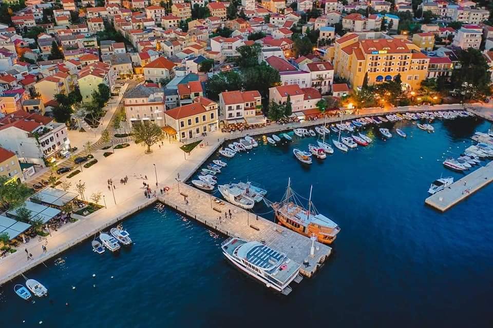 An aerial view captures the vibrant marina, showcasing several boats docked along a well-lit waterfront. The picturesque town features red-tiled roofs, with warm lights illuminating the buildings and pathways. The calm waters reflect the evening glow, enhancing the serene landscape.