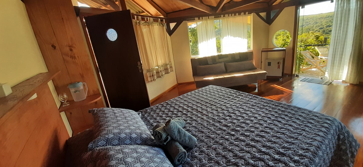 A spacious bedroom features a large bed with a patterned blanket and neatly arranged towels. Natural light filters through sheer curtains, illuminating the wooden interior. A seating area with a comfortable sofa is visible near a glass door, which opens to scenic views of the surrounding nature.