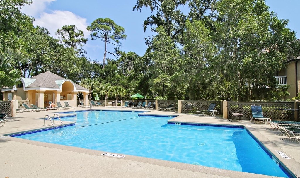 An outdoor pool area is surrounded by lush greenery, with sun loungers placed along the edge. A shallow area is indicated, and a shaded pavilion is visible in the background, offering additional seating and shade for relaxation.