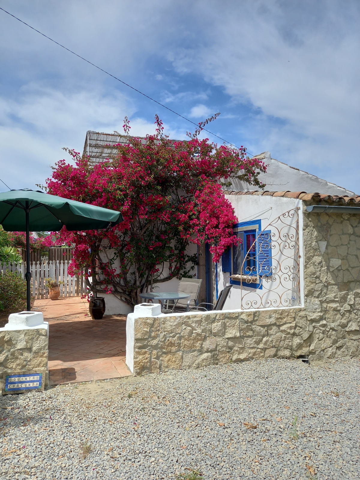 The exterior of the cottage is adorned with vibrant pink bougainvillea, creating a colorful contrast against the white stone walls. A green umbrella shades a small seating area, providing a welcoming spot to relax. The entrance is framed by a rustic stone pathway and decorative ironwork.