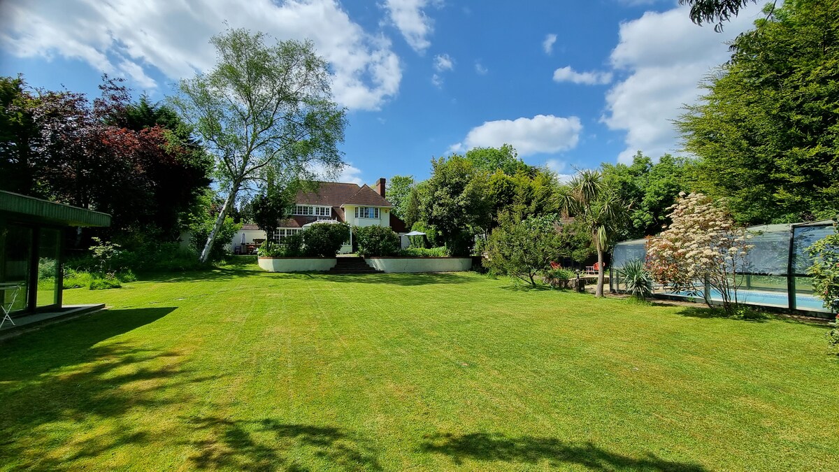 A spacious garden area is surrounded by mature trees and flowering plants, offering a tranquil setting. The guest house is visible in the background, with a well-maintained lawn leading to a sparkling salt-water swimming pool. The sky is partly cloudy, creating a bright atmosphere.