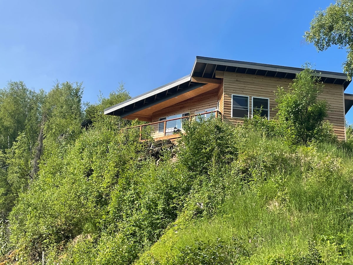 A modern cabin is set atop a hillside, surrounded by lush greenery. Its wooden exterior features large windows and a balcony that offers a scenic view of the landscape. The clear blue sky contrasts with the vibrant surrounding foliage.