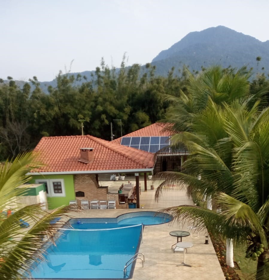 A welcoming outdoor area is seen, featuring a blue swimming pool surrounded by palm trees. A spacious house with a red tiled roof and solar panels is visible alongside, with mountains in the background creating a serene landscape.