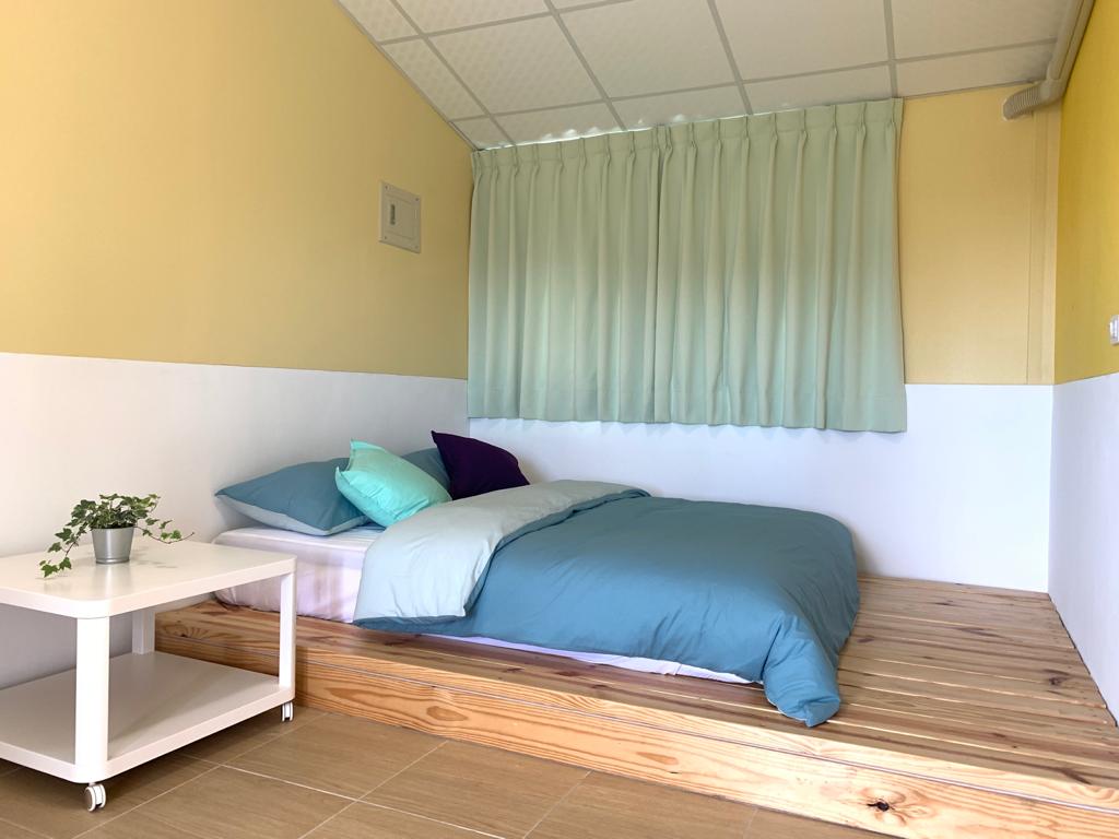 A cozy sleeping area features a wooden platform bed with a soft teal and purple bedding arrangement. A small white side table is positioned nearby, adorned with a potted plant. Bright light filters in through sheer green curtains, enhancing the room's serene atmosphere.