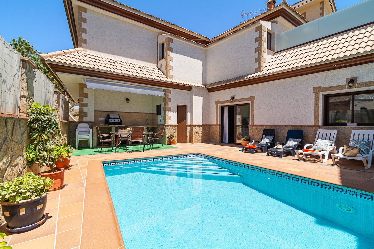 A sunny terrace features an inviting swimming pool surrounded by lounge chairs. A shaded barbecue area is visible, along with a dining set suited for outdoor meals. Lush potted plants add greenery to the space, contributing to a relaxing environment.