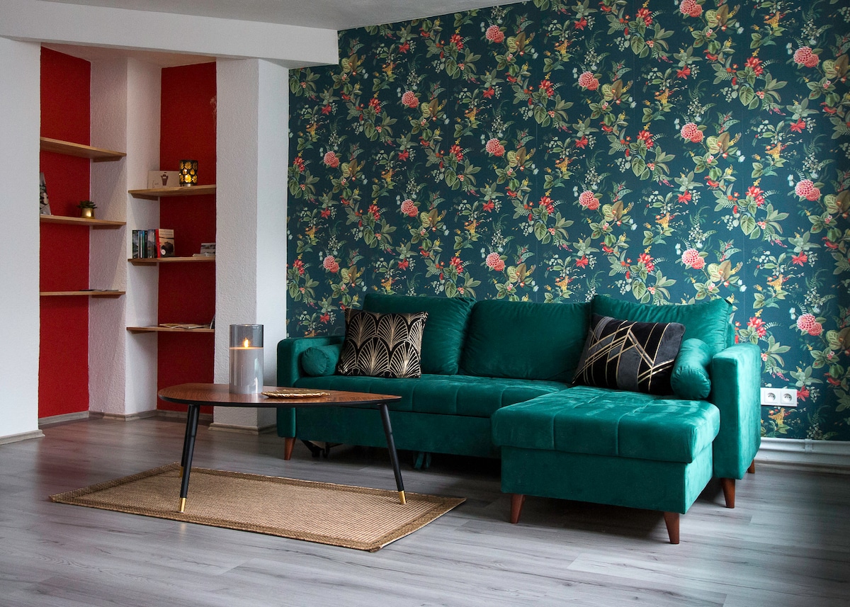 A spacious living area features a green upholstered sofa against a vibrant floral wallpaper. A medium-sized wooden coffee table sits on a light-colored area rug, complemented by decorative pillows on the sofa. A shelf in the background displays various items, adding a personal touch.