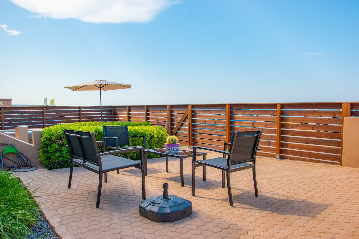 An outdoor seating area is presented with four black metal chairs surrounding a glass-top table. A large umbrella offers shade, while a vibrant green bush adds a touch of nature. The patio features warm, tan paving stones and a wooden fence providing privacy.