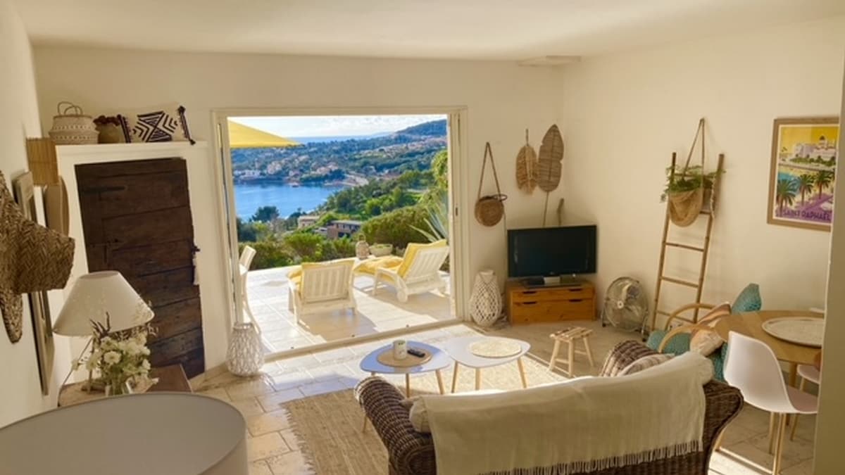 A bright living area opens onto a panoramic view of the sea through large glass doors. Comfortable seating is arranged thoughtfully, complemented by natural decorations. Light colors enhance the airy atmosphere, while outdoor seating is visible on the terrace beyond.
