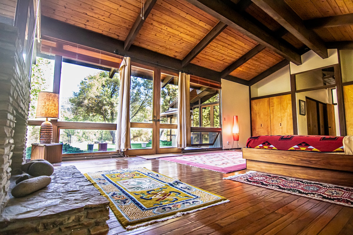 A spacious room features high wooden ceilings and large windows offering views of the surrounding nature. A comfortable bed is positioned against the wall, while decorative rugs add warmth to the hardwood floor. Natural light pours in, highlighting the eclectic decor.