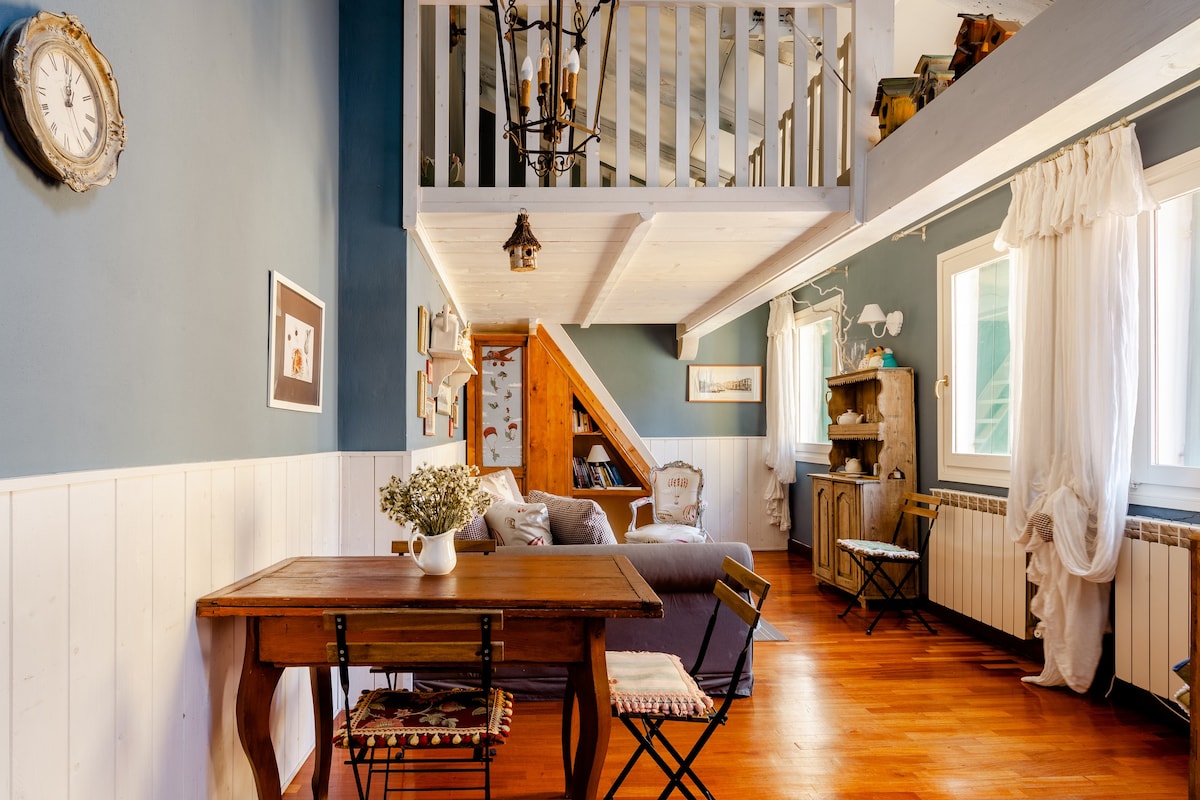 A cozy living space features wood flooring and pale blue walls. A wooden table is positioned near a comfortable sofa, with structured seating arranged thoughtfully. Large windows allow natural light to illuminate the area, and a ladder leads to an upper loft. Decorative plants and artwork are present.