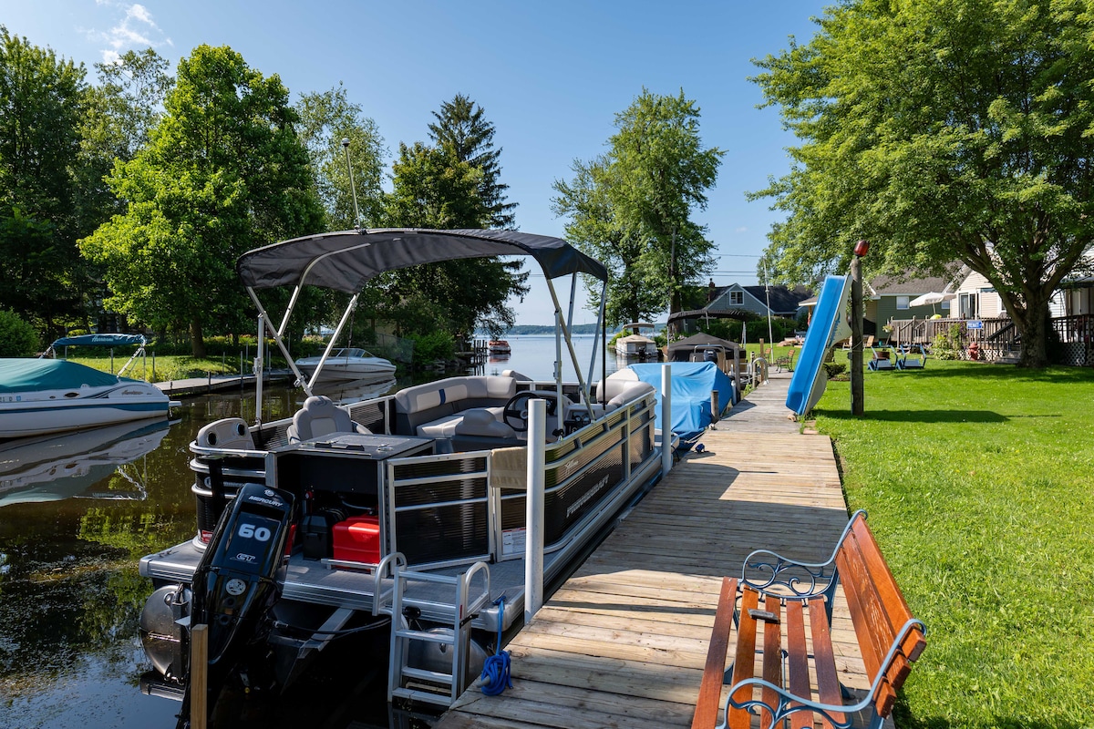 A spacious dock is lined with a variety of boats, including a pontoon with a covered canopy. The surrounding greenery enhances the serene setting, while a wooden bench is positioned nearby for relaxation. The calm waters lead out towards Chautauqua Lake, framed by trees.
