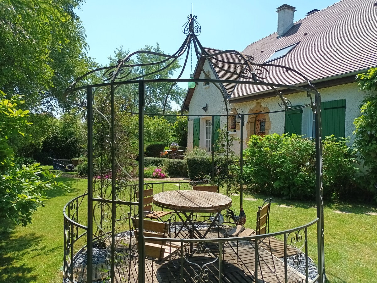 A charming gazebo with intricate wrought iron design is centered on a wooden deck, surrounded by lush greenery in a landscaped garden. A circular table with matching chairs is visible, providing an inviting space for outdoor relaxation. The backdrop includes a house with green shutters.