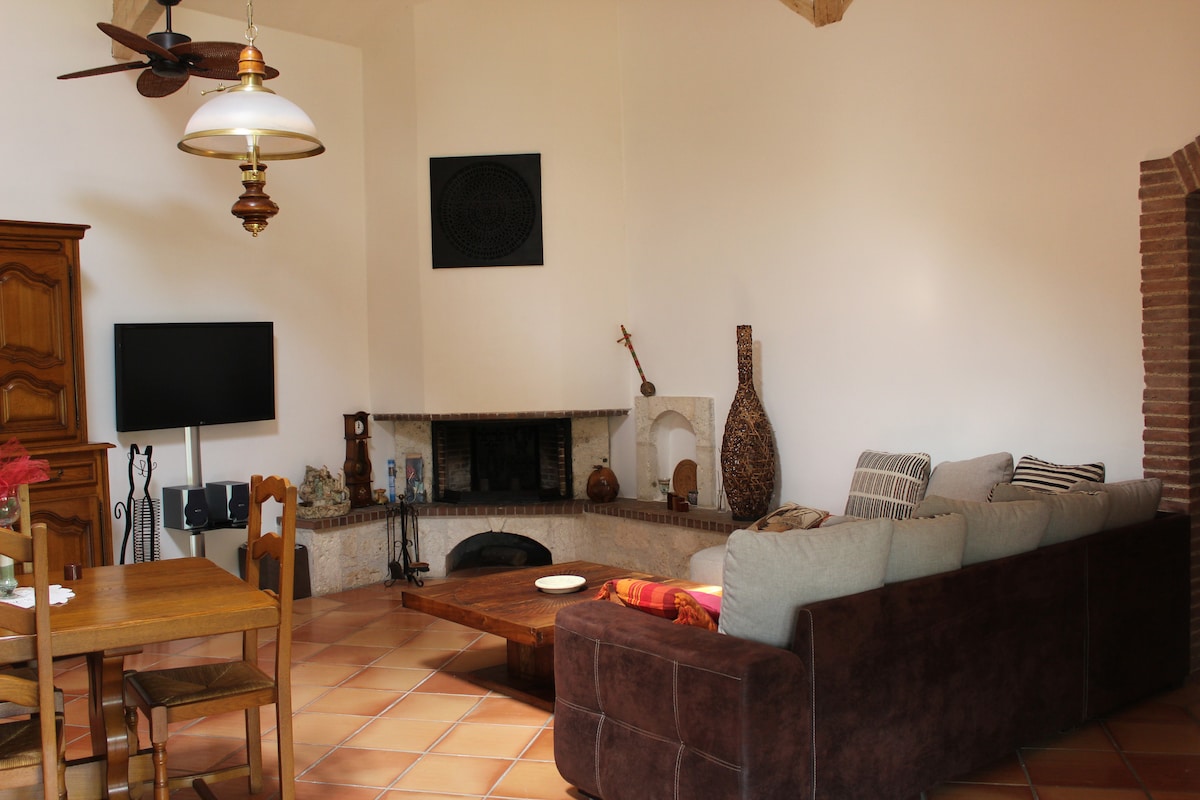 A cozy living area features a spacious sectional sofa and a large coffee table, complemented by a rustic wooden dining set. A flat-screen television is mounted on the wall, while a fireplace adds warmth to the space. Natural light enters through the windows, enhancing the inviting ambiance.