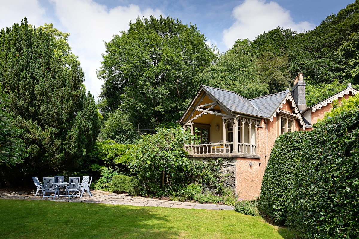 The exterior of the house is framed by lush greenery, with a spacious terrace adorned with outdoor seating. The garden area, featuring well-maintained lawns and hedges, creates a serene environment. The charming architectural details of the house invite guests to enjoy the natural surroundings.
