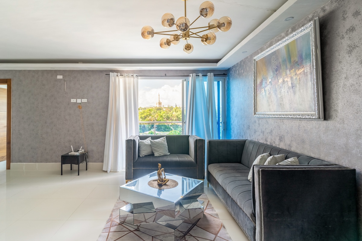 A stylish living area features a gray velvet sectional sofa and a modern glass coffee table. Large windows allow natural light to fill the space and offer a view of the outdoors. Decorative accents include soft cushions and a framed painting on the wall.