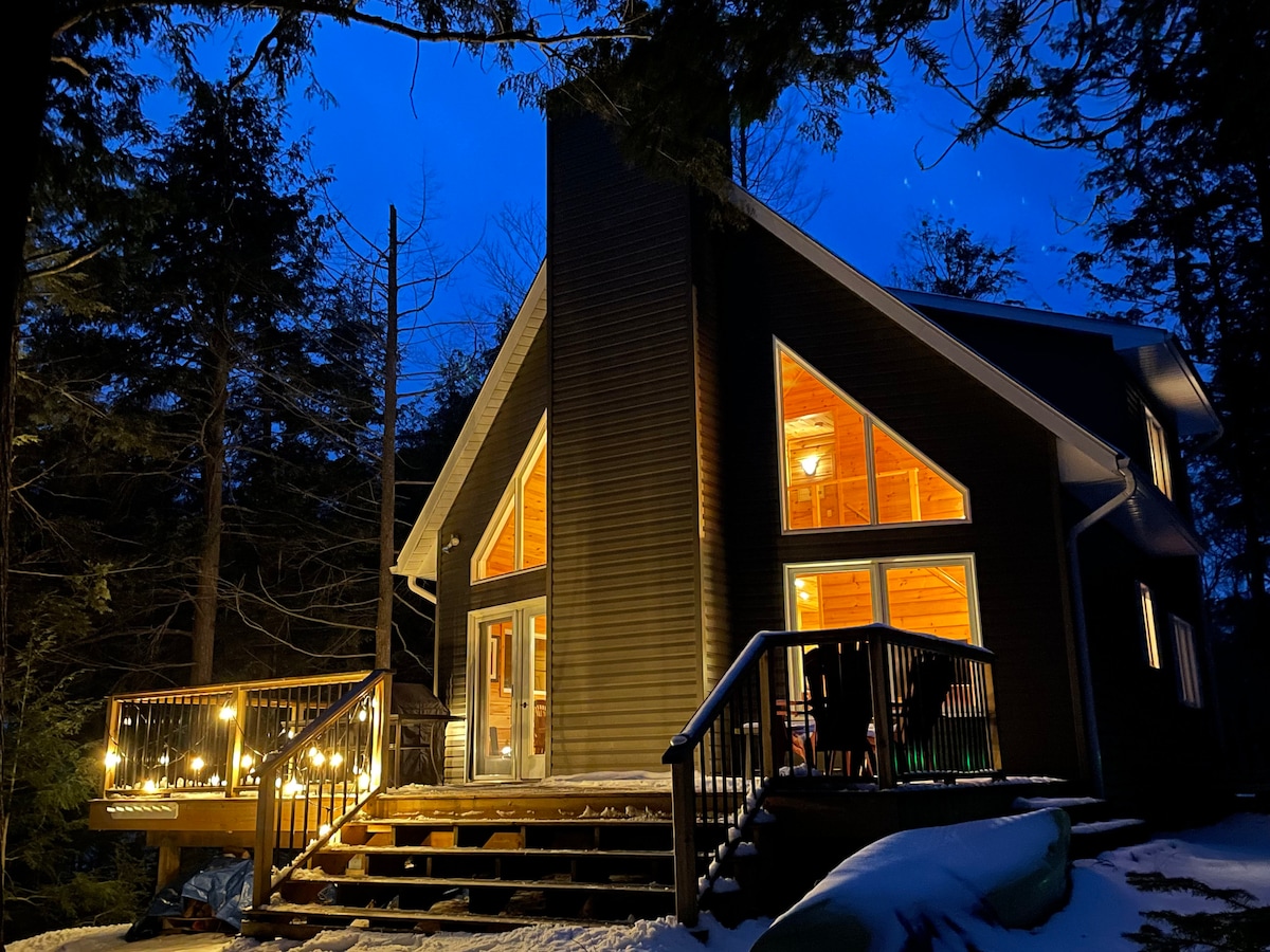The exterior of the cottage is illuminated at night, showcasing triangular windows reflecting a warm glow. A spacious deck is visible, equipped with seating and decorative lights. Surrounding trees create a natural setting, enhancing the peaceful ambiance of the property.