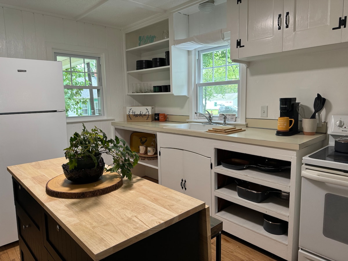 A bright kitchen is equipped with white cabinets and a light-colored countertop. An island features a wooden top with a potted plant. Natural light enters through a window above the sink, while a coffee maker and a range add functionality to the space.