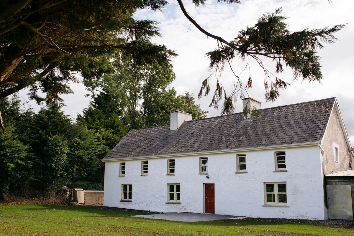 A historic two-story house with a white exterior stands amidst a serene countryside setting. Large windows line the front, allowing natural light to fill the interior. A sloped roof is complemented by a welcoming red door. Surrounding greenery enhances the tranquil atmosphere.